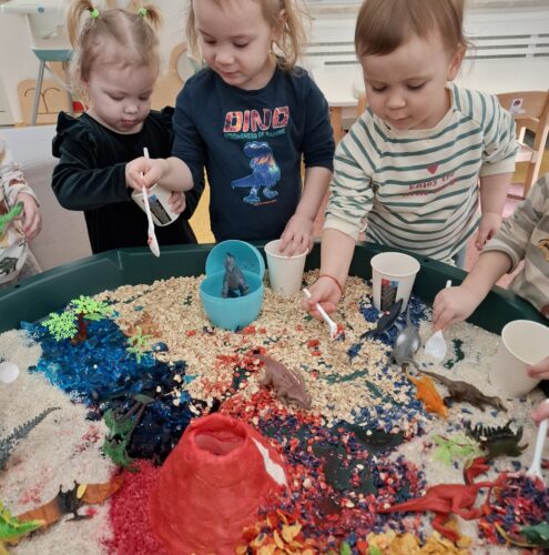 dzieci bawią się na planszy sensorycznej z dinozurami - zabawkami i przesypują makraon itp